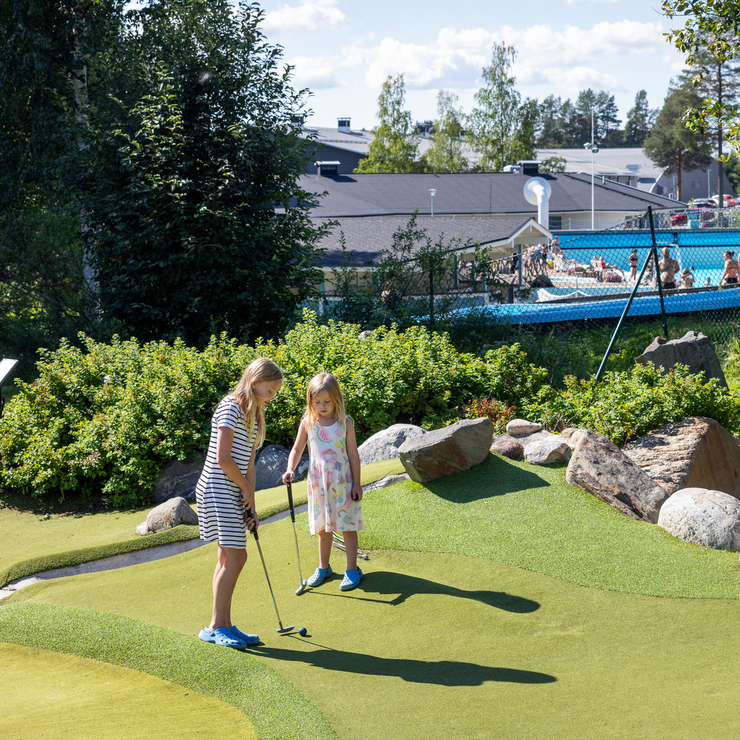 Två flickor spelar minigolf på campingens äventyrsgolf. De är sommarklädda i shorts och t-shirt. I bakgrunden syns badet