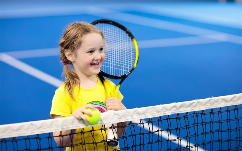 Flicka i gula träningskläder som står med sitt racket vid ett tennisnät. Banan i bakgrunden har en klar blå färg.
