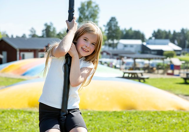 Flicka klättrar i en klätterställning på en lekplats. I bakgrunden syns stora hoppkuddar och picknickbord.