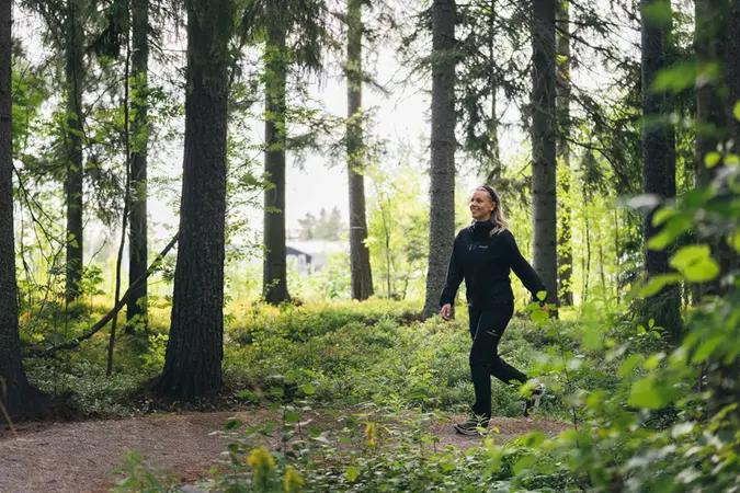 En kvinna vandrar i skogen. Vandringsleden är en skogsstig, stora tallar ramar in leden från båda sidorna.