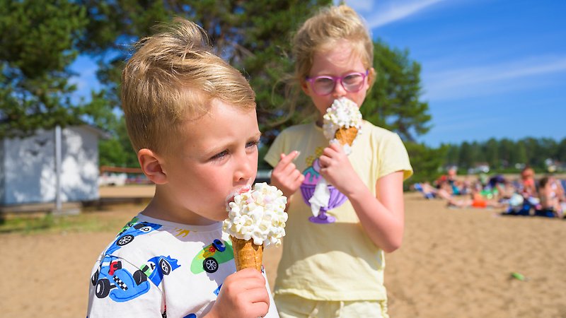 Två barn står på stranden och äter var sin mjukglass ur strut.