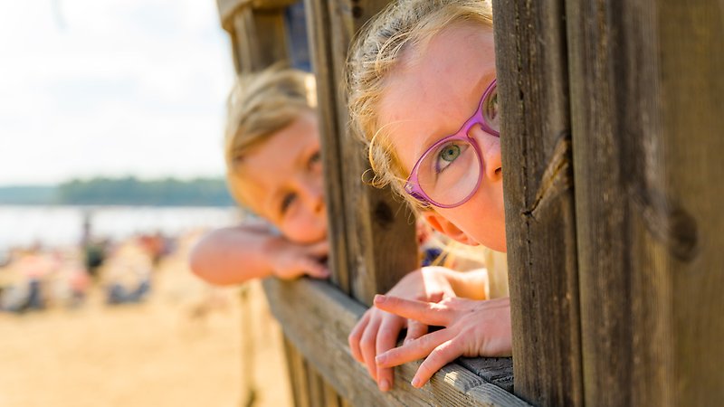 Två barn tittar ut genom varsin glugg i träskeppet som finns att leka i på stranden.
